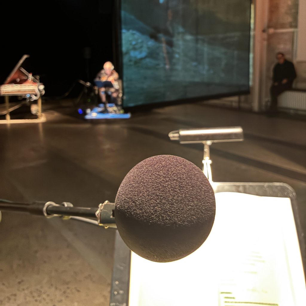 Im Vordergrund ein Mikrofon und ein Textblatt. Im Hintergrund verschwommen ein Musiker, ein Cembalo und eine Leinwand.
